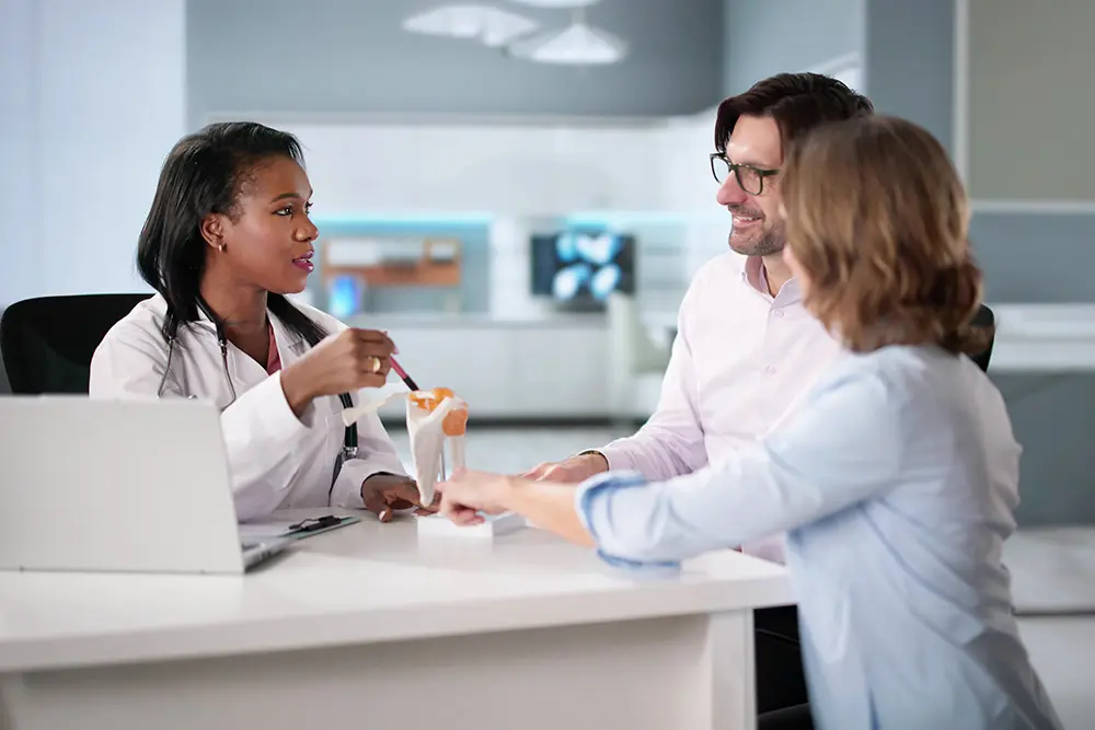 A female doctor explains a medical model of a knee joint to a smiling man and woman across a desk in a modern office setting. The doctor gestures with a pen, and a laptop sits on the desk.