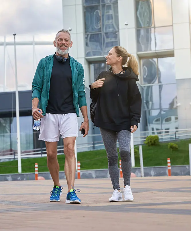 An older man and woman dressed in athletic wear are walking and smiling outdoors in an urban area, with a modern glass building in the background. The man holds a water bottle and has headphones around his neck.